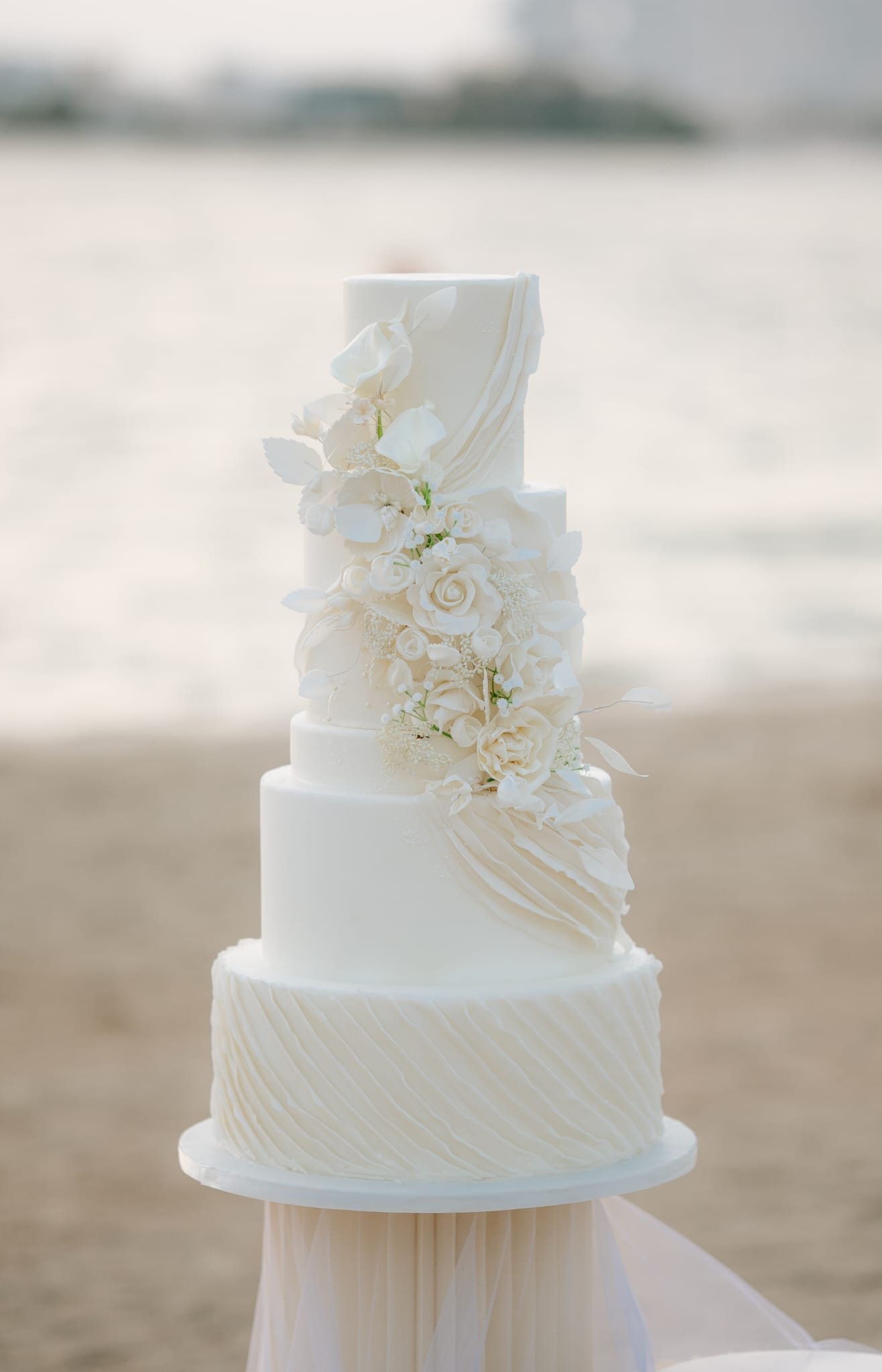 A tall white wedding cake with sugar flowers on a beach at sunset