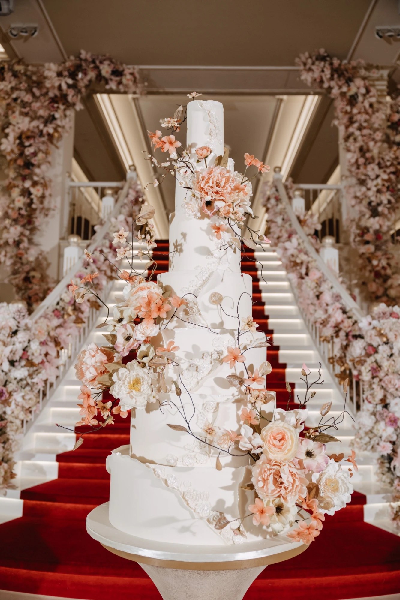 A tall white wedding cake with peach and cream sugar flowers on a floral-draped staircase with a red carpet