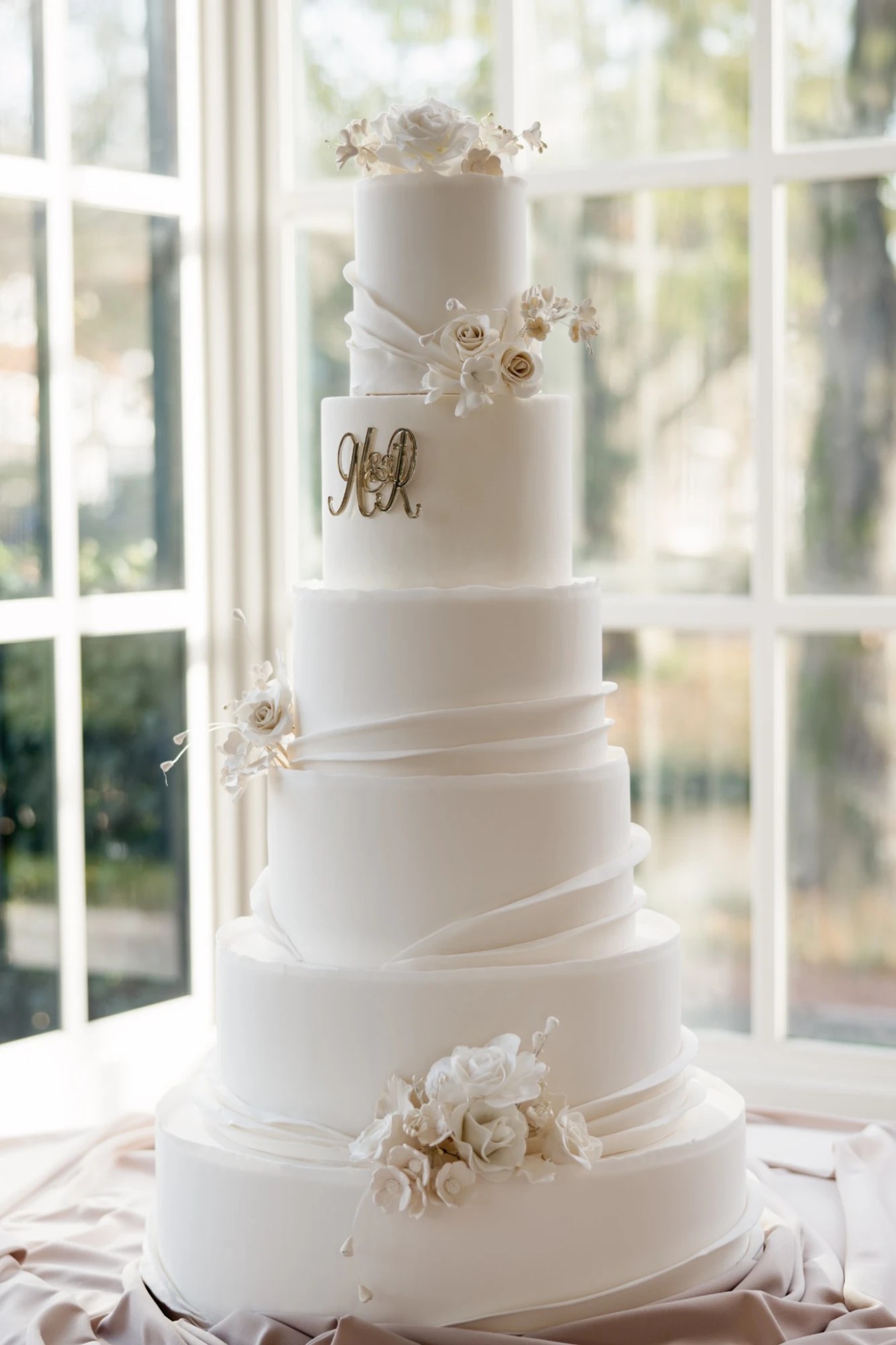 A six-tier white wedding cake with draped fondant, a gold monogram and ivory sugar roses beside tall windows