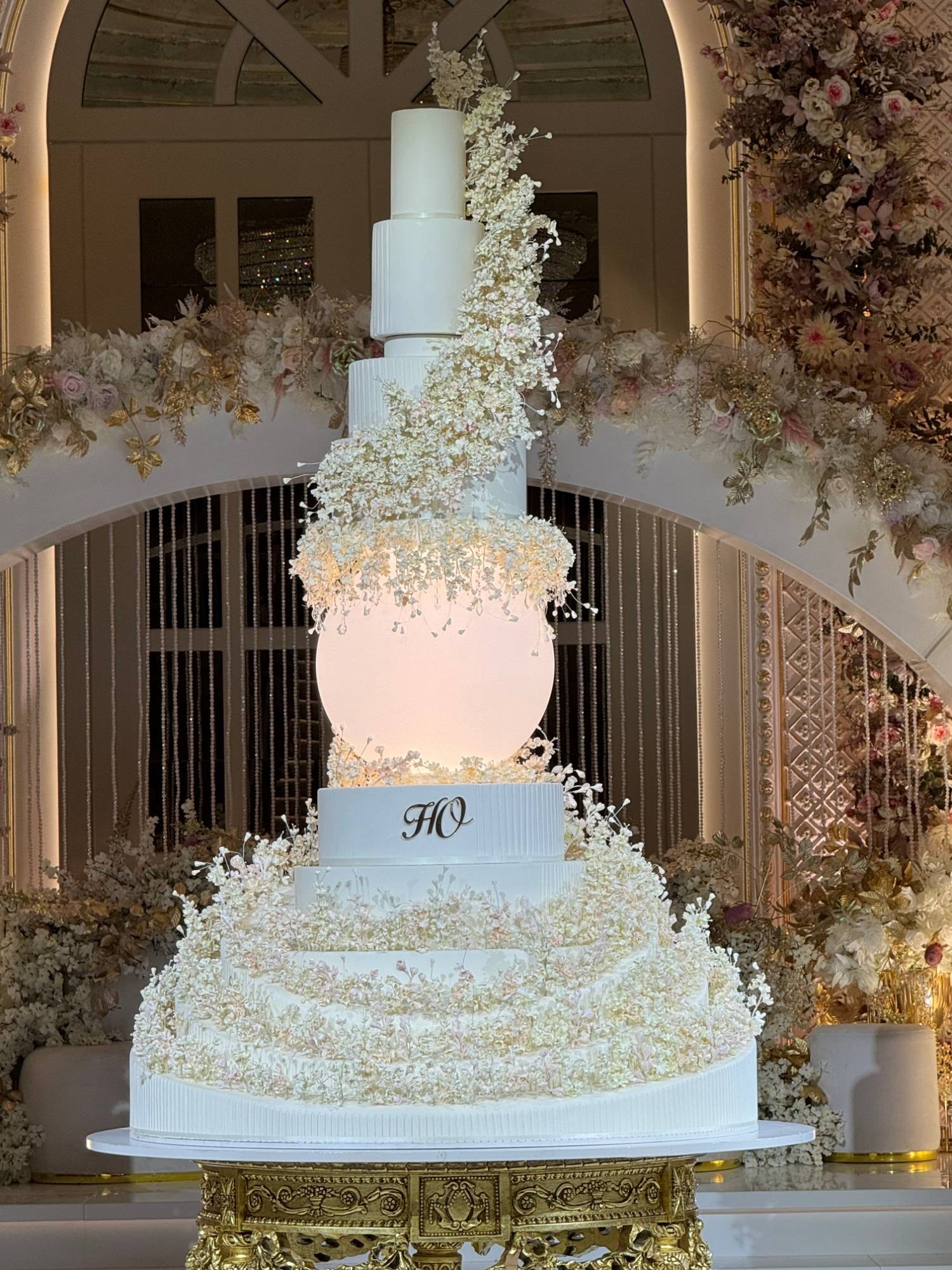 A tall white wedding cake with a glowing illuminated sphere between its tiers and cascading ivory sugar blossoms, before a floral arch