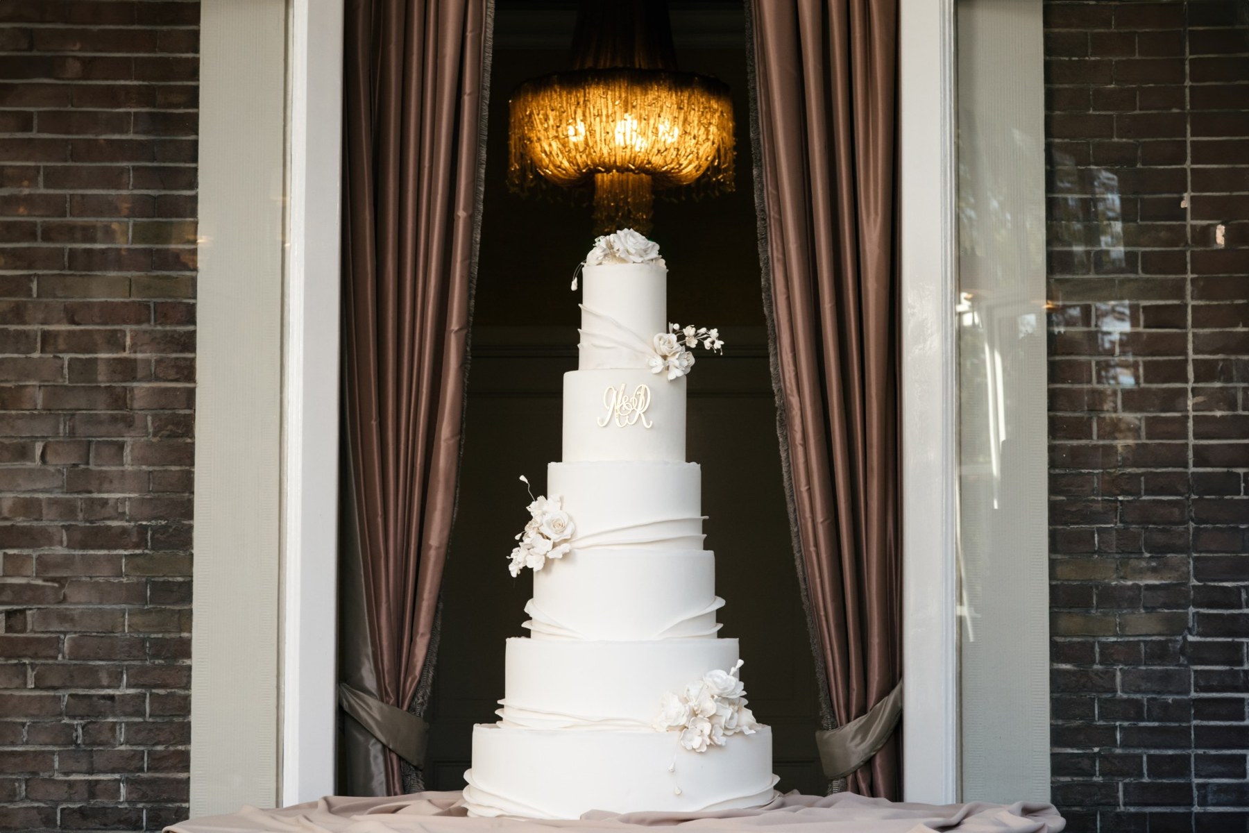 A six-tier white wedding cake with fondant drapes, a monogram and ivory sugar flowers framed by brown curtains and a chandelier