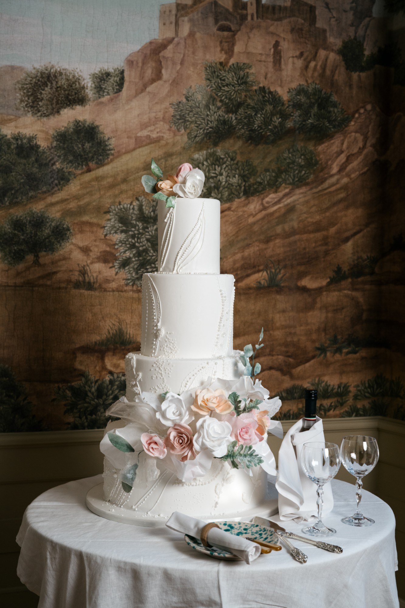 A four-tier white wedding cake with pink, peach and white sugar roses and hand-piped detail, on a linen-covered table